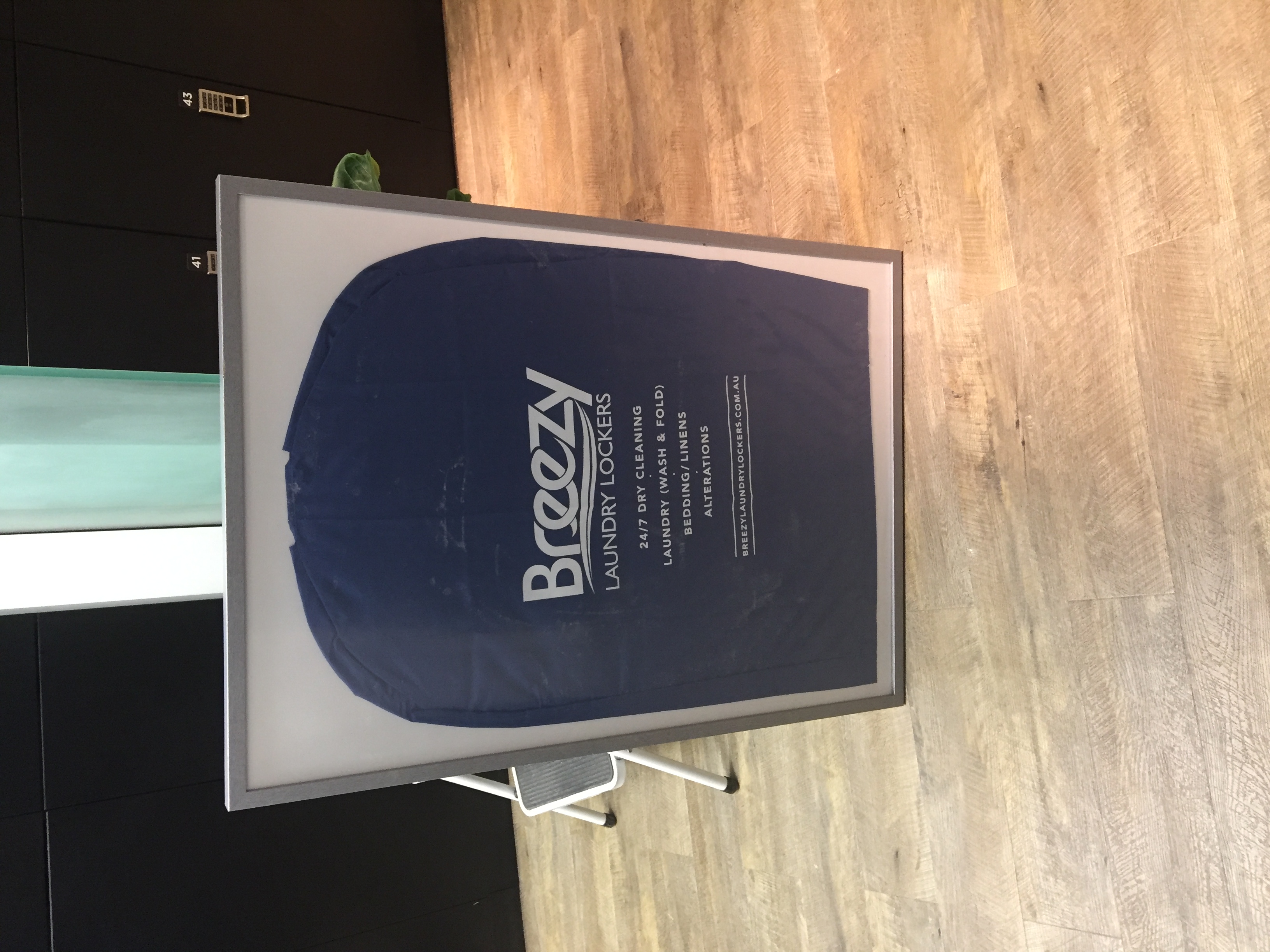 Breezy Laundry Lockers branded poster on an easel listing the 24/7 services available — dry cleaning, wash and fold, bedding, alterations