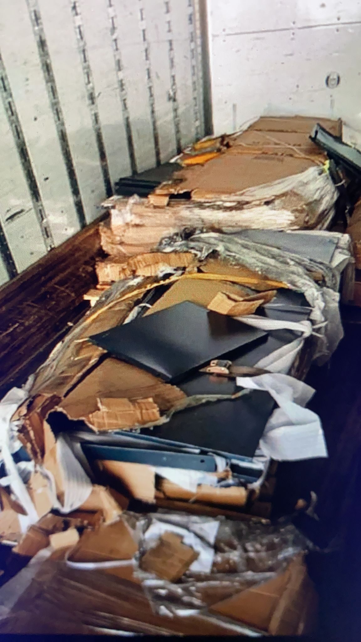 Inside a freight truck with damaged Breezy laundry locker components. Black metal locker panels exposed through ripped cardboard packaging after international shipping.