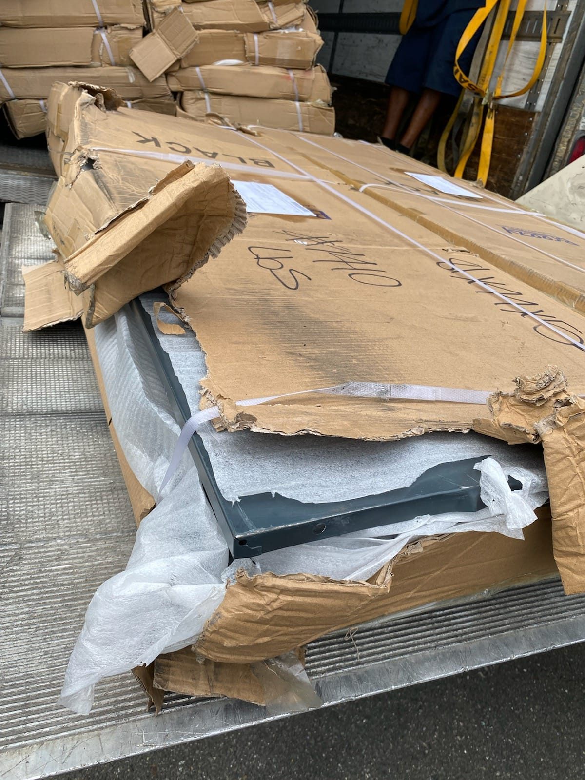 Close up of damaged cardboard packaging showing the bare metal frame of a Breezy laundry locker exposed through torn box walls and split strapping after long-haul freight.