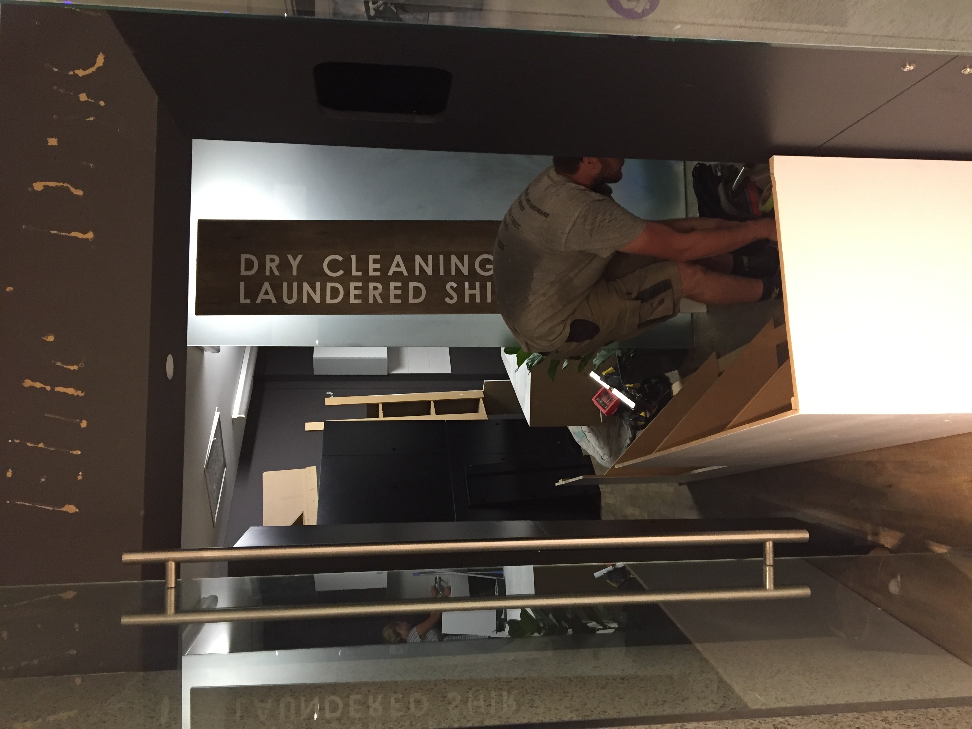 Worker bending over inside the half-built Elite Dry Cleaners store, installing fixtures with the Dry Cleaning and Laundered Shirts wood signage already mounted