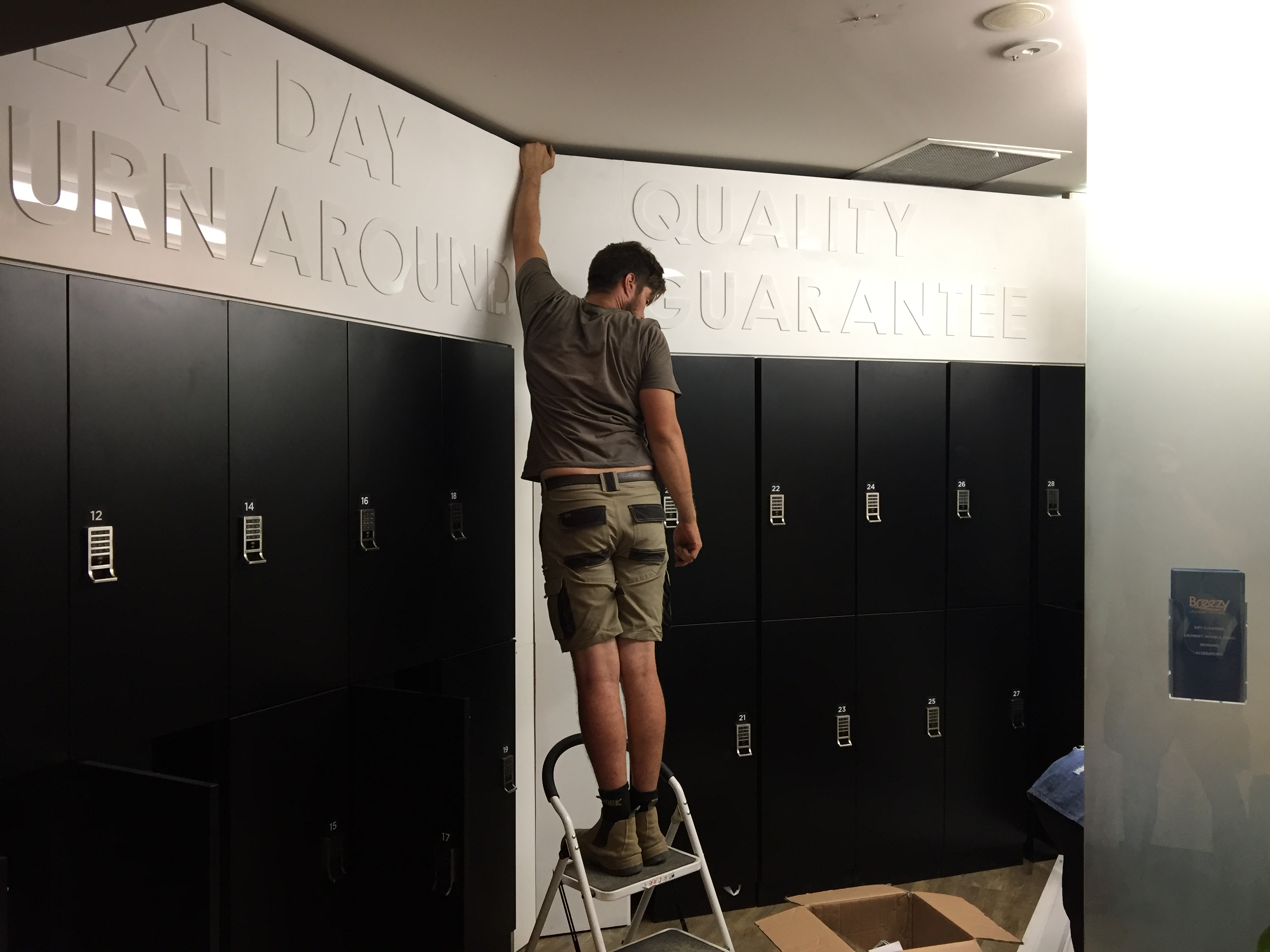 Worker on a stepladder installing the Next Day Turn Around and Quality Guarantee cut-out wall lettering above the locker bank